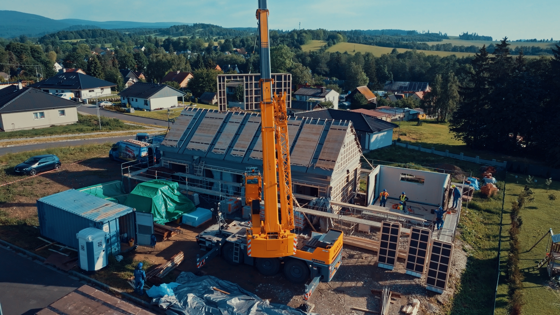 Crane placing prefabricated timber house panels, with containers and materials nearby